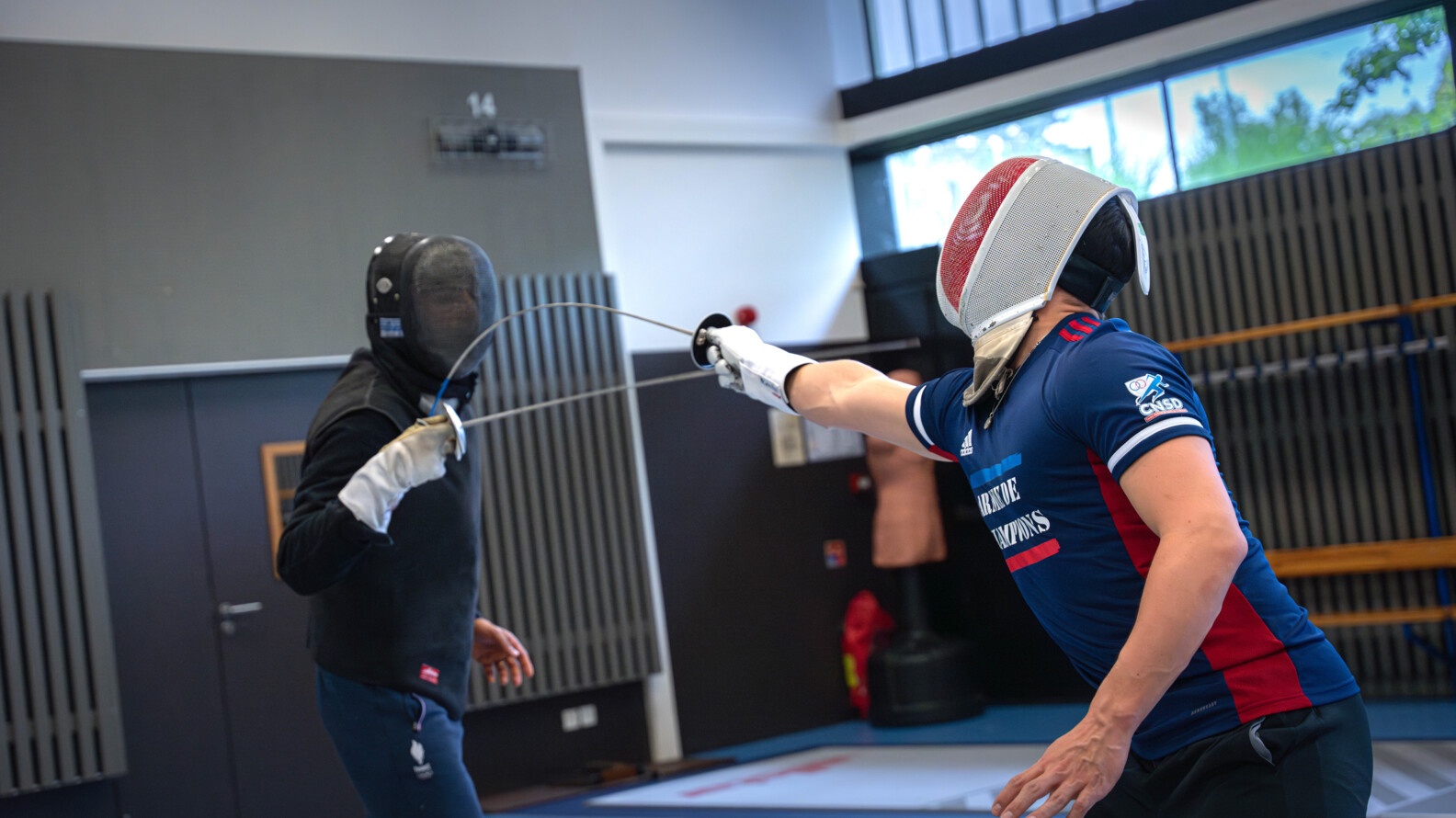 A fencer dressed in the tracksuit of the army of champions touches his fencing master in front of him, dressed in black, who does not parry