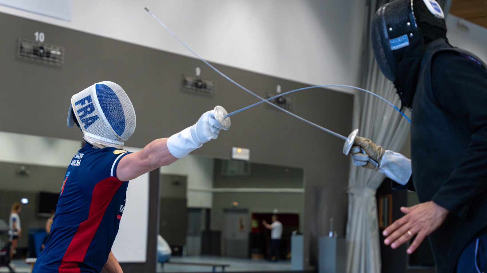 Two fencers confront each other, helmeted, one on the right dressed in black and the second, dressed in blue and red, touching his opponent with his foil whose tip is covered in blue