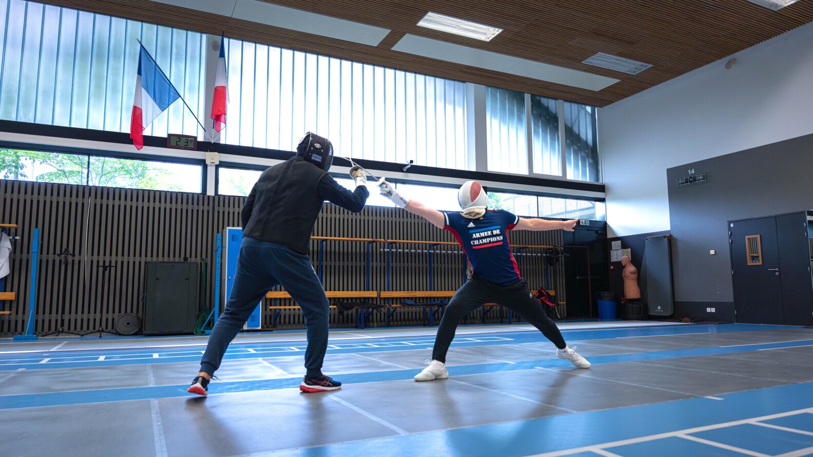 Profile view of two fencers, on the left he is dressed in black and makes a parade facing his opponent, dressed in blue and red, who extends to touch him