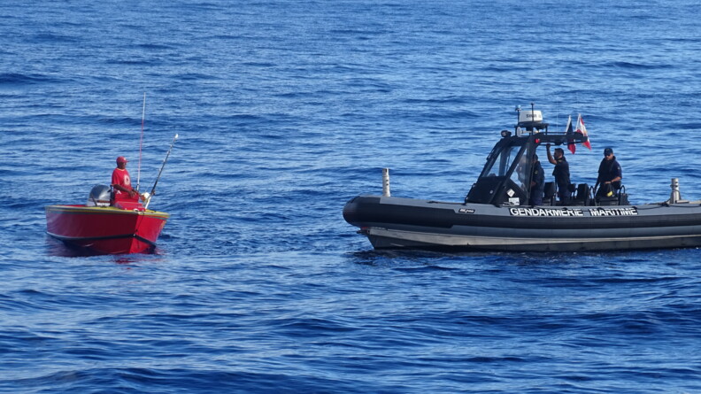 Photo d'un zodiac noir portant sur son flanc l'inscription gendarmerie maritime, avec quatre gandarmes à son bord, s'approchant d'une petite embarcation rouge, sur laquelle se tient debout un pêcheur, vêtu d'un t-shirt et d'une casquette rouge.