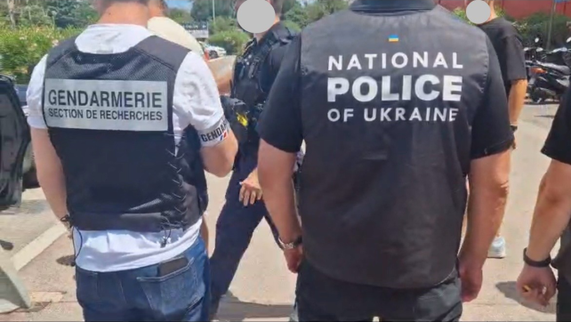 De dos à gauche, un enquêteur en civil portant au bras droit un brassard gendarmerie et une chasuble noire "gendarmerie section de recherches" et à droite, de dos, un enquêteur arborant une chasuble noire "national police of ukraine"