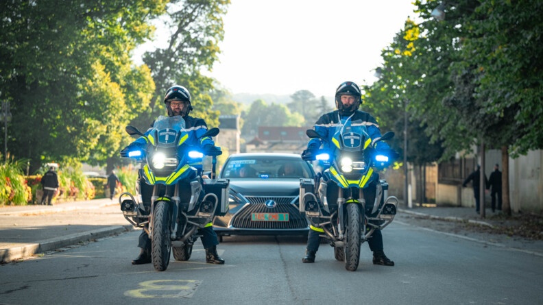 Des les rues du centre ville de Villers-Cotterêts, deux gendarmes motocyclistes précèdent une berline de couleur sombre. Les gyrophares bleus de chacun des trois véhicules sont allumés.