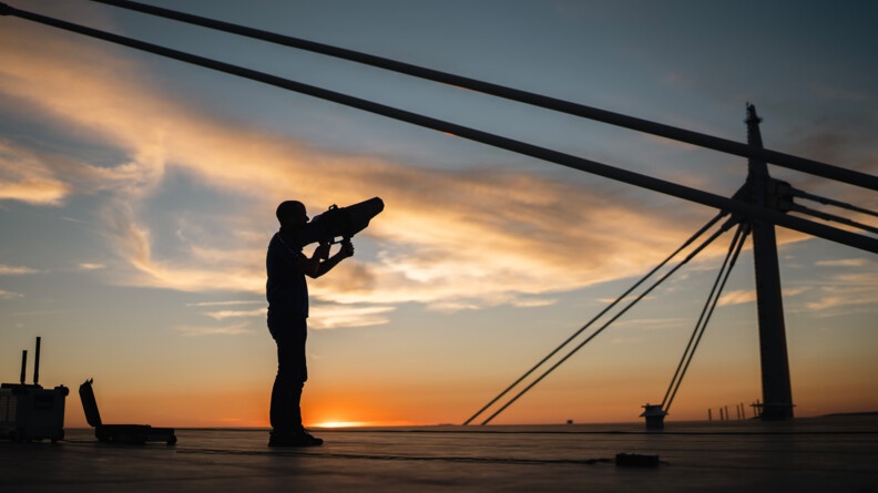 Sur le toit du Stade de France, un homme se tient debout, de biais, face au ciel où le soleil se couche, un fusil anti-drone dans les mains.