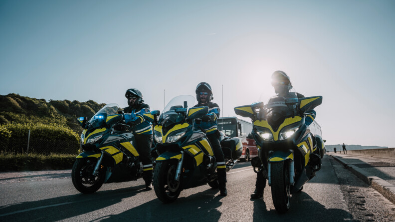 Trois motards de la gendarmerie sur une route, pris en contre plongée avec derrière un grand ciel bleu et le soleil.