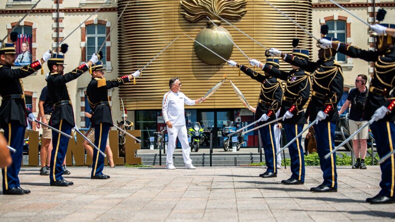 Devant le musée de la gendarmerie nationale, un homme vêtu de blanc passe la flamme olympique à un autre relayeur, sous la voûte d'acier de huit éléèves-offieirs de la gendarmerie en tenue de tradition