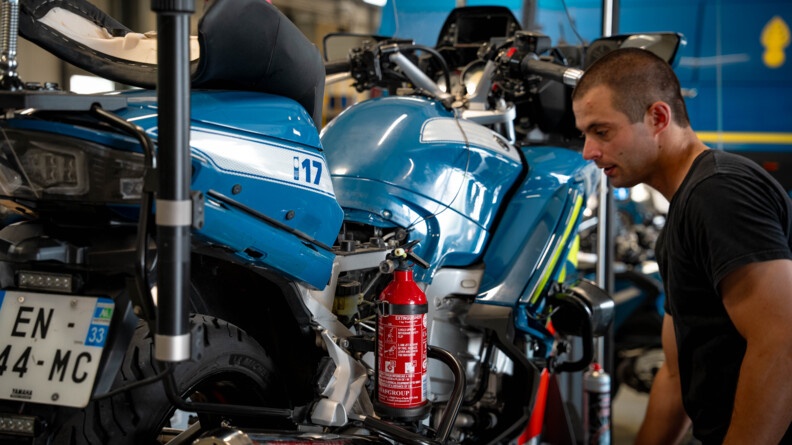 Dans un atelier de réparation aitomobile, un mécanicien se tient debout, à côté d'une moto bleue de la gendarmerie. L'homme est vêtu d'un t-shirt et d'un pantalon noirs.