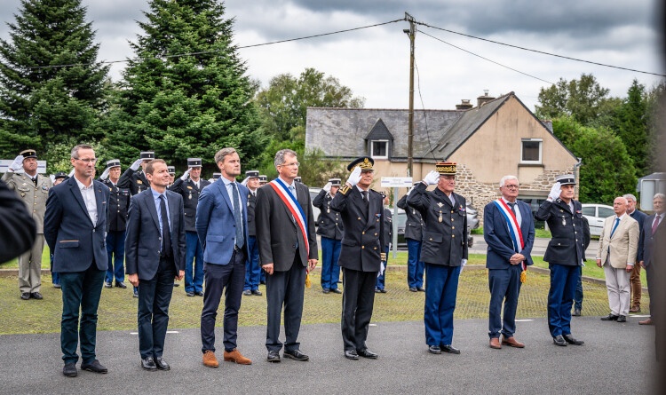 Huit personnes, soit trois élus, trois militaires (effectuant un salut) et trois civils, se tiennent debout côte à côte, dans un village. Autour d'eux sont réunis d'autres militaires ainsi que des civils.