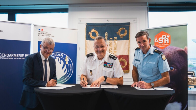 Devant des kakemonos de la gendarmerie et de la SPA, derrière de hautes tables rondes recouvertes de tissu noir, à gauche un homme en costume blue marine prteur de luenttes, au centre un général 5 étoiles de la gendarmerie en chemisette blanche, à droite un général trois étoiles de la gendarmerie en chemisette bleue, tous trois tiennet un stylo en main et s'aprrêtent à signer un document posé sur les tables