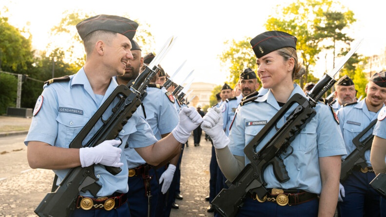 Deux gendarmes d'un des deux Régiments d'infanterie de la Garde républicaine se font un "tcheck" de la main, souriants, face à la caméra, ils se regardent, avec l'Arc de triomphe en arrière plan