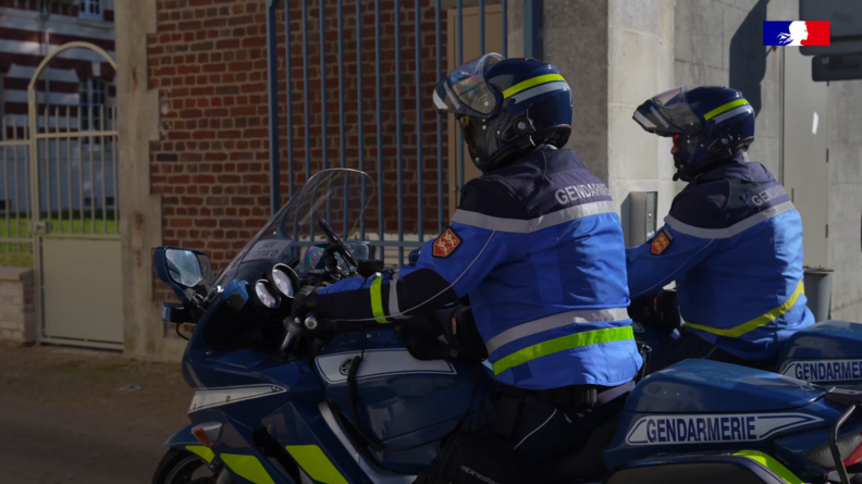 Deux motards de la gendarmerie, au guidon de leur moto, que l'on voit de dos, pénètrent dans l'enceinte d'un bâtiment en briques rouges