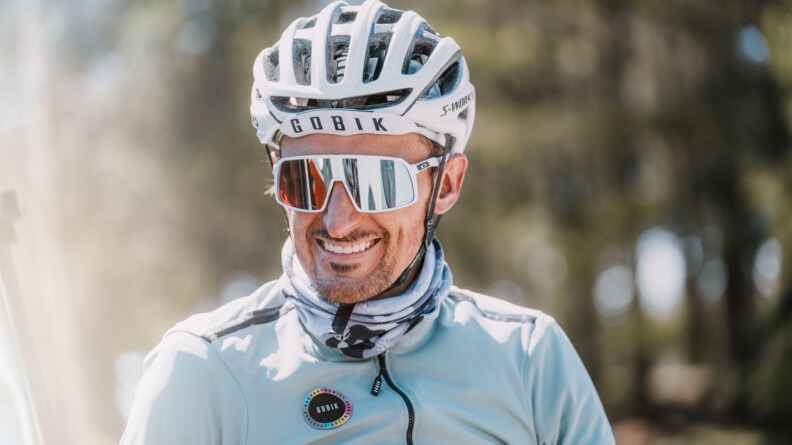 Un cycliste, dont on aperçoit uniquement le haut du corps, regarde l'objectif en souriant. Il es vêtu d'un blouson bleu ciel, d'un casque blanc et de lunettes de soleil blanches.