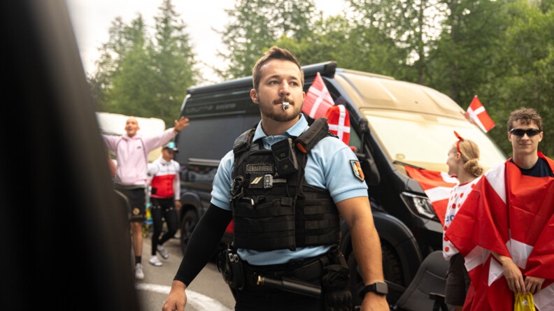 Un gendarme se tient debout sur la route, au milieu de la foule, sifflet dans la bouche.  Derrière lui, on aperçoit une camionnette évoluant sur la route.