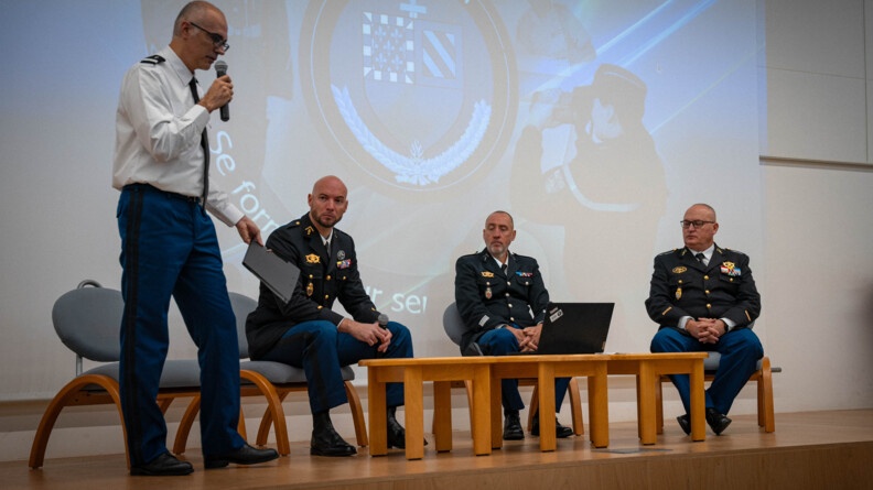 Quatre militaires se tiennent sur une estrade. L'un d'eux est debout, portant un micro à sa bouche. Les trois autres sont assis côte à côte, écoutant le gendarme se trouvant debout. Derrière eux,  un grand écran projette un écusson sur lequel est inscrit "Ecole de Dijon"