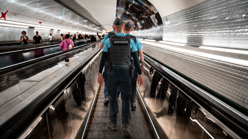 Trois gendarmes de dos remontent un couloir de métro carrelé de blanc, sur un tapis roulant. A gauche, deux autres tapis roulants où l'on apercoit des usagers dans le flou, le premier va dans le même sens que les gendarmes, le second en sens inverse.