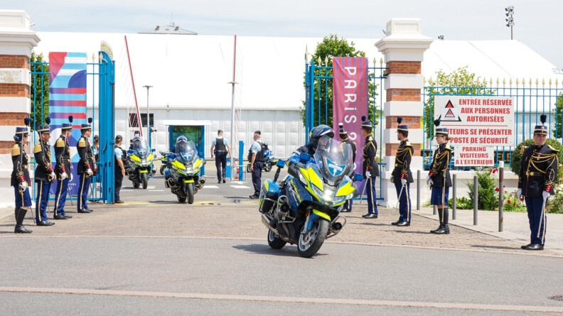 Trois motard de la gendarmerie sortent par le portail d'une grille bleue aux couleurs des Jeux de Paris 2024. De part et d'autre, huit élèves-officiers en tenue de tradition