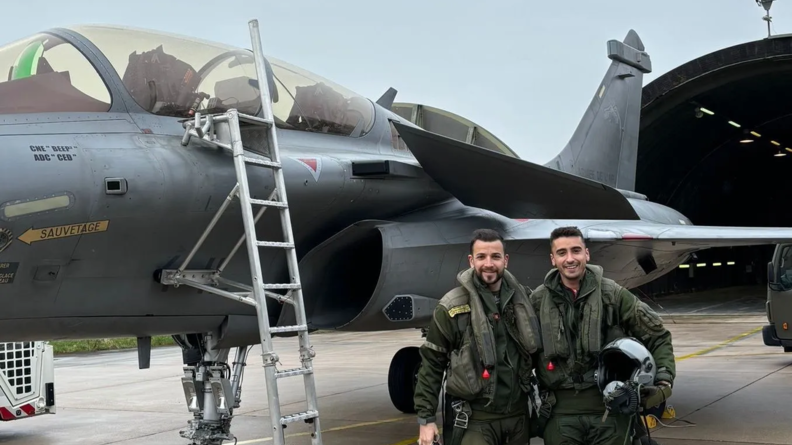 Deux pilotes en tenue kakie se tiennent côte à côte, souriants, casque à la main, devant un avion de chasse Rafale, sur le tarmac.