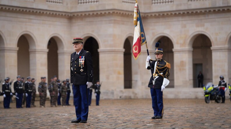 Le directeur général de la gendarmerie au garde-à-vous au milieu de la cour d'honneur de l'Hôtel national des Invalides. Derrière lui, un gendarme en grande tenue porte le drapeau tricolore de la gendarmerie nationale. Dans le fond à gauche, une partie des troupes au garde-à-vous ; à droite, un motocycliste. En arrière-plan, les arches de la cour d'honneur.