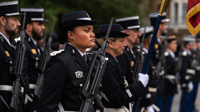 Des militaires en uniforme se tiennent de debout lors d'une cérémonie officielle, leur fusil contre la poitrine. En premier plan, se trouve une femme, que l'on voit de biais