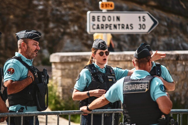 Quatre gendarmes, un de profil, une femme de face, en train de parler et d'indiquer une direction, un homme de dos, la main contre une barrière, et un quatrière que l'on devine. Les quatre militaires sont sur le bord d'une route dans le Vaucluse.