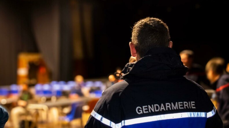 Un gendarme vêtu d'un blouson gendarmerie, vu de dos, dans une salle. En arrière-plan on devine des tables et des chaises, et à droite trois pompiers.