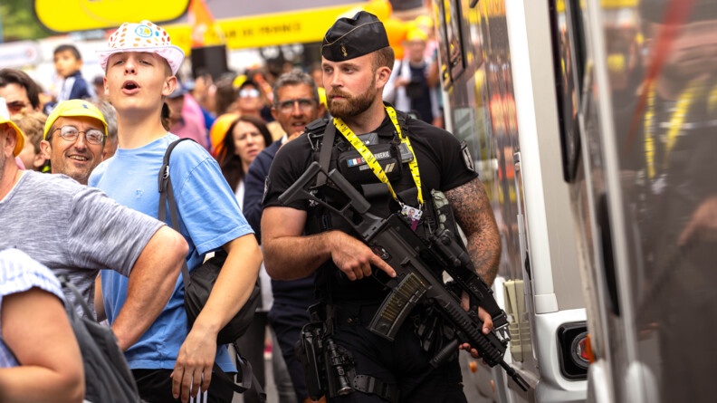 Un gendarme, revêtant un uniforme noir, se tient debout sur la route, au milieu de la foule. Il tient dans ses mains un fusil.