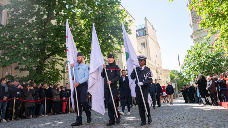 Au centre de la cour de l'hôtel de Beauvau, un gendarme, une sapeur pompiers et une policière avancent fièrement en portant les drapeaux des JO de Paris.