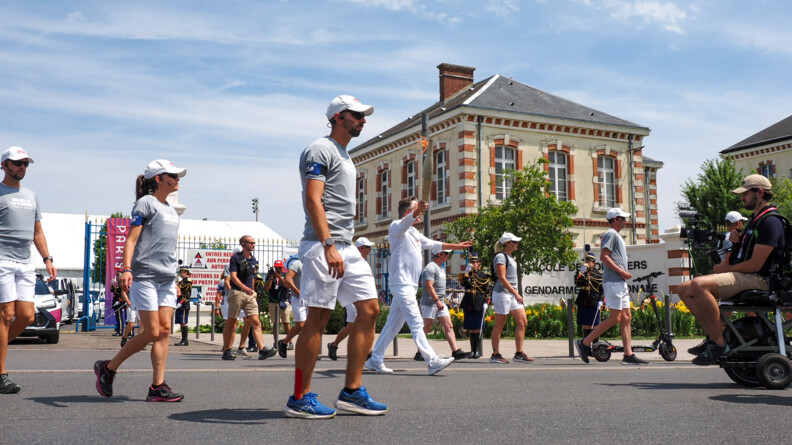 Un homme vêtu de blanc portant la flamme olympique entourés d'une dizaine d'hommes et de femmes en polo gros et short blanc, casquette blanche et micro portatif, devant des bâtimens anciens