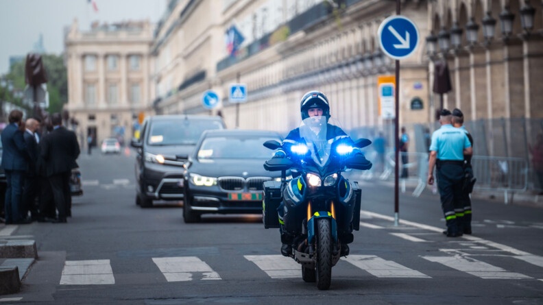 Une moto de la gendarmerie roule dans une rue de Paris, phares bleus allumés à l'avant, devant une voiture noire dotée d'une plaque d'immatriculation verte. Sur la chaussée, est écrit, en grandes lettres blanches : PARIS.