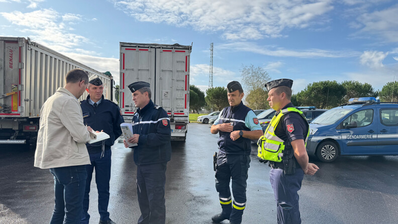 4 gendarmes et un civil sur un parking où se trouvent des camions en arrière-plan et des voitures à droite