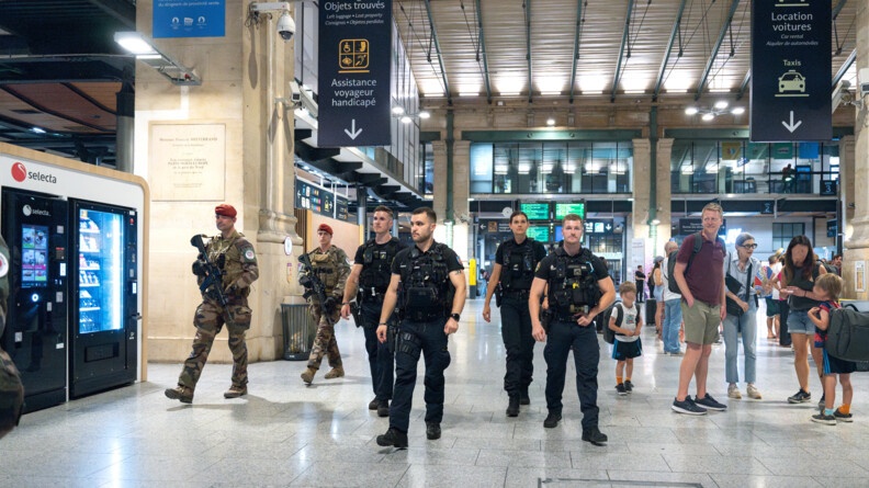 Quatre gendarmes mobiles de face, patrouillent dans un hall de gare. A gauche, deux militaires de Sentinelle en treillis patrouillent à leurs côtés. A droite, un groupe de voyageurs.