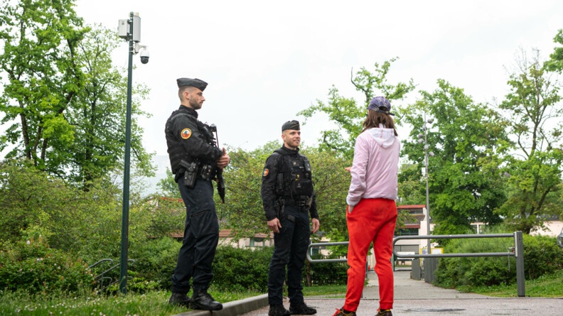 Deux gendarmes discutent avec un adolescent portant une casquette violette, un blouson rose, un jogging rouge et des baskets. lls se tiennent debout, dans l'espace extérieur et verdoyant d'une résidence