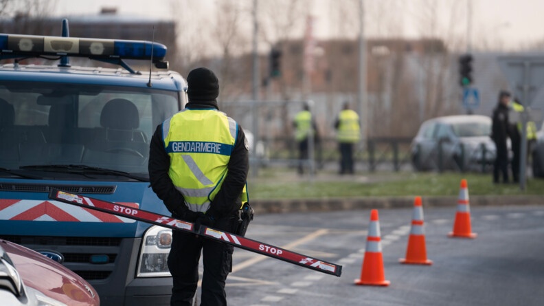 Un gendarme de dos portant un stop stick devant un véhicule de la gendarmerie. Sur la route, des plots orange. en arrière-plan, d'autres gendarmes