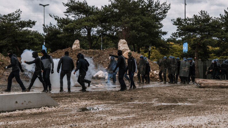 Une douzaine de plastrons repoussés par les gendarmes mobiles dans un exercice sur un terrain boueux. De la fumée de lacrymogne monte du sol.