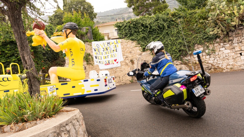 Une gendarme motocycliste roule sur la route, dans un virage, à la suite d'un véhicule jaune LCL. La gendarme porte une tenue bleue et un casque blanc, dont s'échappe une tresse blonde.