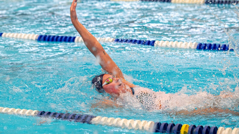 Une jeune femme nage dans un bassin, crawlant sur le dos. Elle porte un bonnet de bain bleu marine et des lunettes de nage.