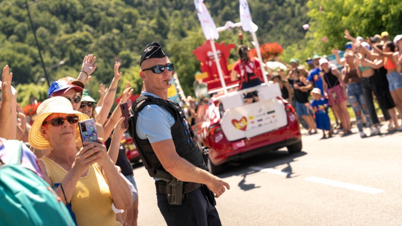 Un gendarme se tient debout, au milieu de la foule, le sifflet dans la bouche, équipé d'un gilet pare-balles et de lunettes de soleil, alors que passent sur la route les véhicules de la caravane du Tour de France 2024. Le temps est ensoleillé et les nombreux spectacteurs présents sur le bord de la route portent pour la plupart un chapeau.