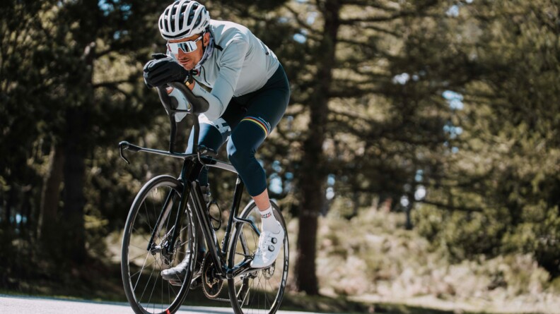 Un cycliste avance sur son vélo, le corps penché en avant, gravissant une côte. Vêtu d'une tenue bleue, il porte un casque, des lunettes de soleil blanches et des gants noirs.