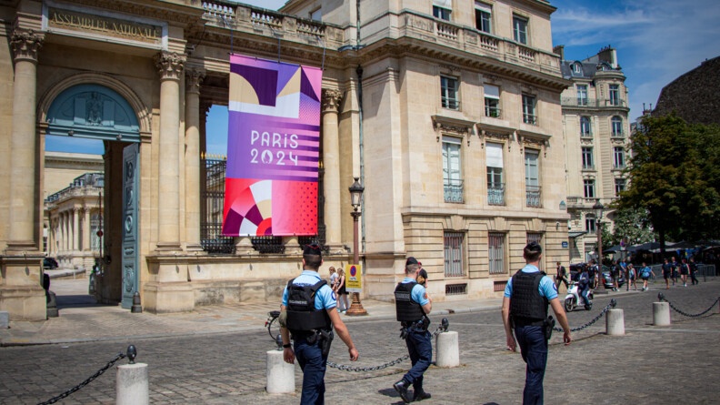 Trois gardes républicains en tenue d'intervention marchant devant un bâtiment Assemblée nationale floquée d'un tissu de Paris 2024