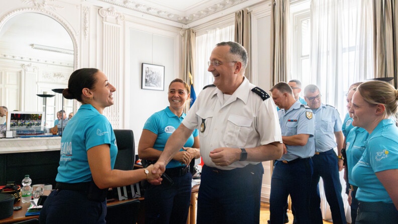 Dans un grand bureau haut de plafond, avec des moulures au plafond et un parquet ancien, le directeur général de la gendarmerie nationale serre la main d'une jeune femme gendarme, vêtue d'un polo bleu turquoise. Dans le dos de cette dernière est inscrite la mention TEAM GENDARMERIE.  A leurs côtés se trouvent d'autres gendarmes.