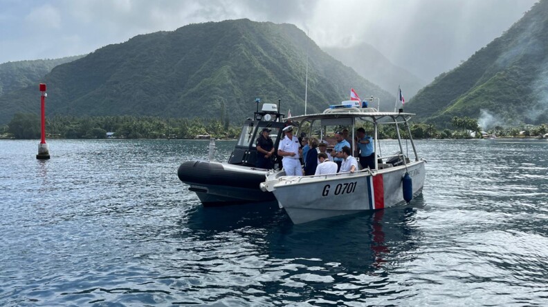 Photo d'une embarcation grise, avec sur la coque des bandes bleues, blanches et rouges, staionnée à côté d'un zodiac noir. Ls deux bâteaux comptent plusieurs personnes à leur bord, dont la plupart se tiennent debout. derrière eux se dressent des reliefs verdoyants. Le ciel est gris et nuageux.