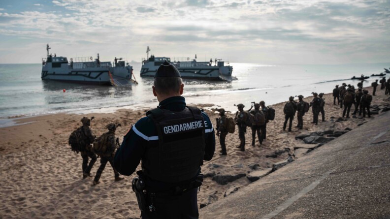 Un gendarme de dos regarde la plage où vient de se dérouler la reconstitution du débarquement. En fond, on observe deux bateaux ainsi que des militaires en tenue.
