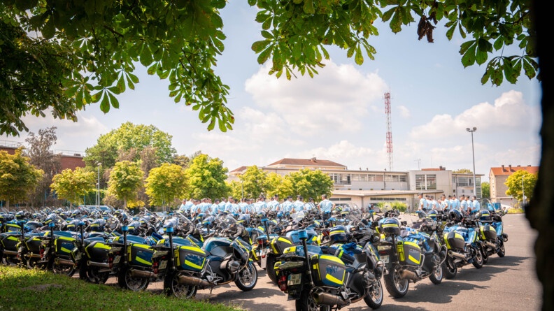 De mombreuses motocyclettes de la gendarmerie sont garées les unes à côté des autres dans une vaste cour. on aperçoit en arrière plan de nombreux gendarmes qui sont réunis.