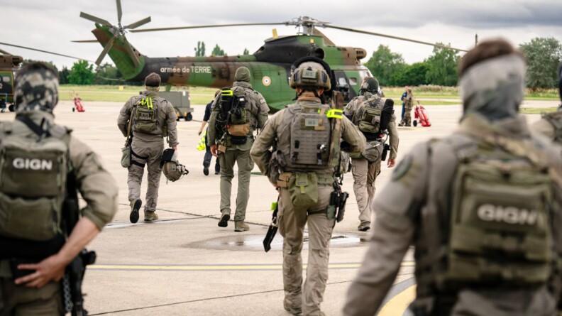 Sept militaires du GIGN en tenue kaki, équipés de casques, armes et gilets pare-balles sont sur le tarmac, en train de se diriger vers un hélicoptère Puma kaki.