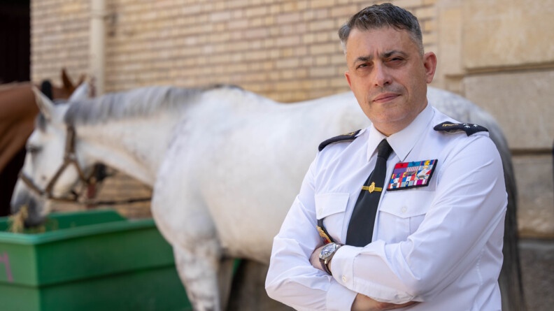 Un général trois étoiles en tenue d'état-major et chemise blanche en buste, les bras croisés. Derrière lui, flou, un cheval blanc en train de manger du foin dans une remorque verte