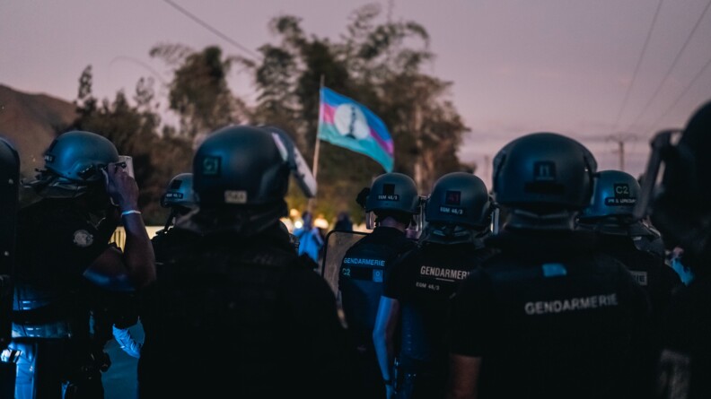 Une dizaine de gendarmes mobiles, en t-shirt et pantalon d'intervention noirs, casque sur la tête, vus de dos. Face à eux, quelques manifestants, dont l'un brandit un drapeau.