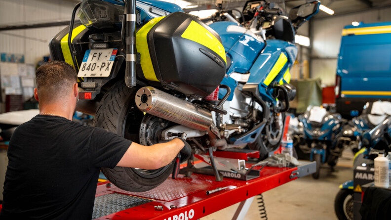 Un homme vêtu d'un t-shirt noir, et de gants noirs, se tient debout de dos, en premier plan. Il répare une moto de la gendarmerie positionnée sur un élévateur. L'homme se trouve dans un atelier de réparation automobile.