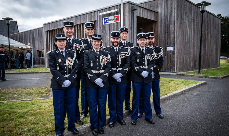 Huit gendarmes en uniforme se tiennent debout, regroupés, devant un bâtiment en bois, sur lequel on apercoit une pancarte aux couleurs bleu, blanc, rouge, portant l'inscription Gendarmerie.