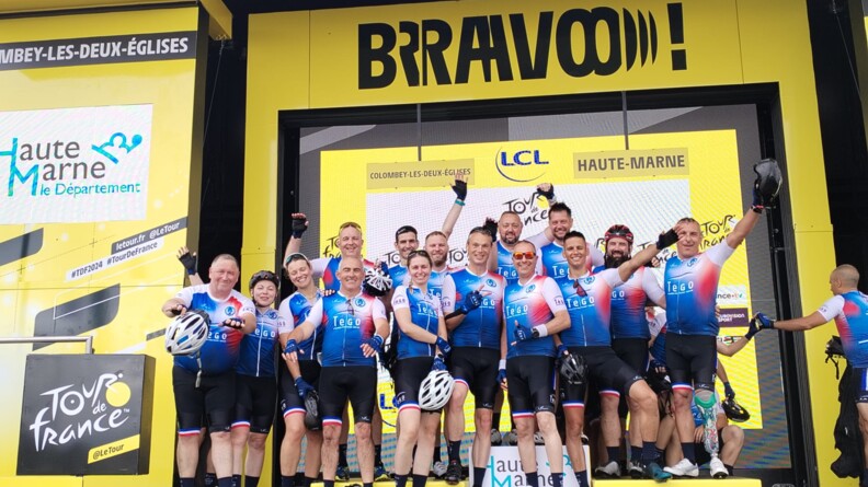 Une quinzaine de gendarmes en tenue de vélo, sur une estrade, faisant des signes de joie,. Le fond est un mur jaune du Tour de France recouvert d'inscriptions de sponsors