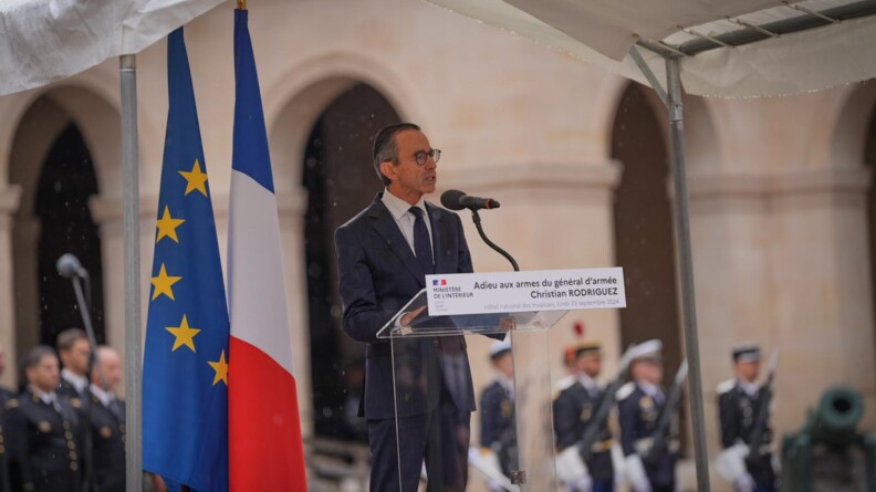 Allocution du ministre de l'Intérieur, debout derrière un pupitre transparent, sous un chapiteau blanc. A sa gauche, le drapeau français jouxte le drapeau européen.