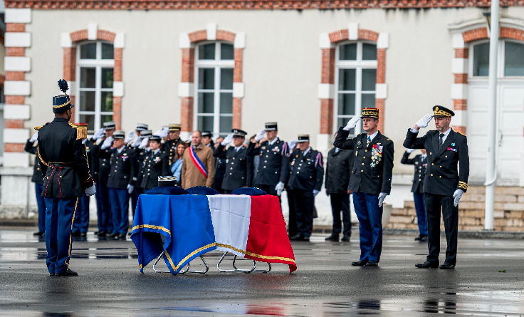 Des militaires saluent un cercueil recouvert du drapeau français. Au milieu d'eux, se trouve un élu, vêtu d'un manteau beige et de l'écharpe tricolore